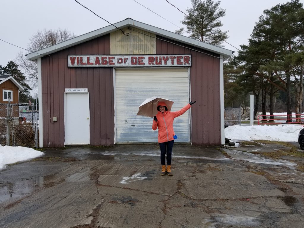 Lerner Fellow Jordana Gilman poses with an umbrella, outside under the Village of DeRuyter sign 